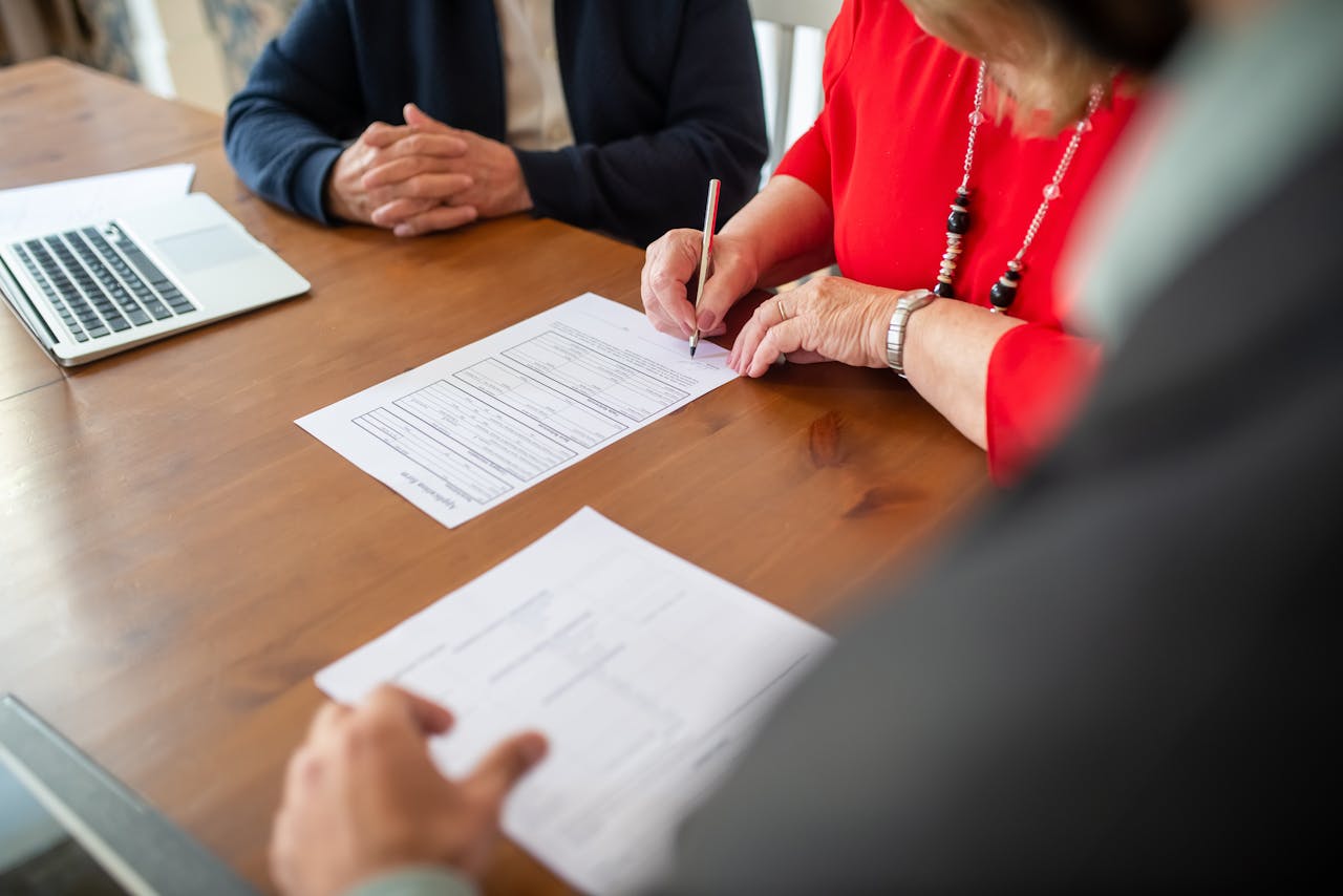 Two adults sign documents on a wooden table at a business meeting, emphasizing teamwork.